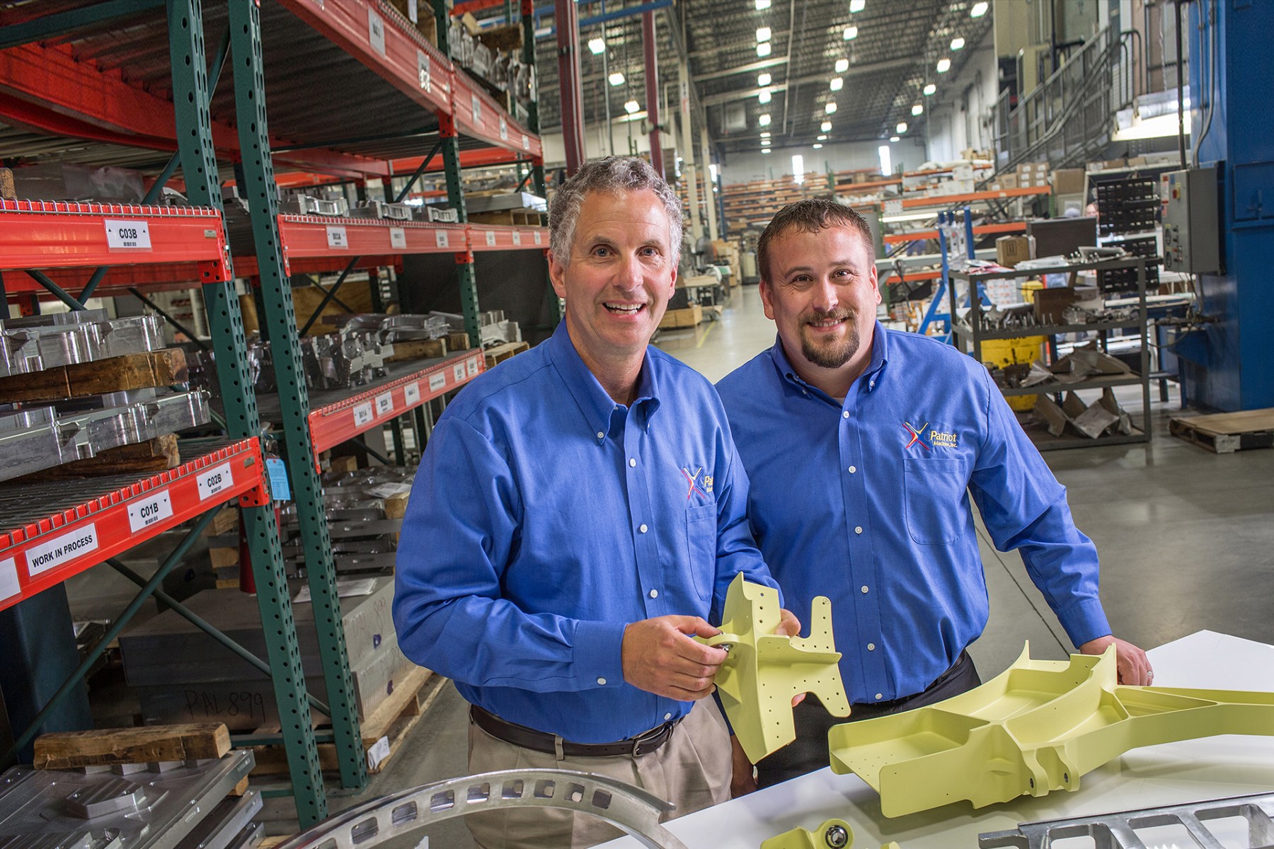Two Patriot Machine associates on the shop floor, with some machined parts on a table in front.
