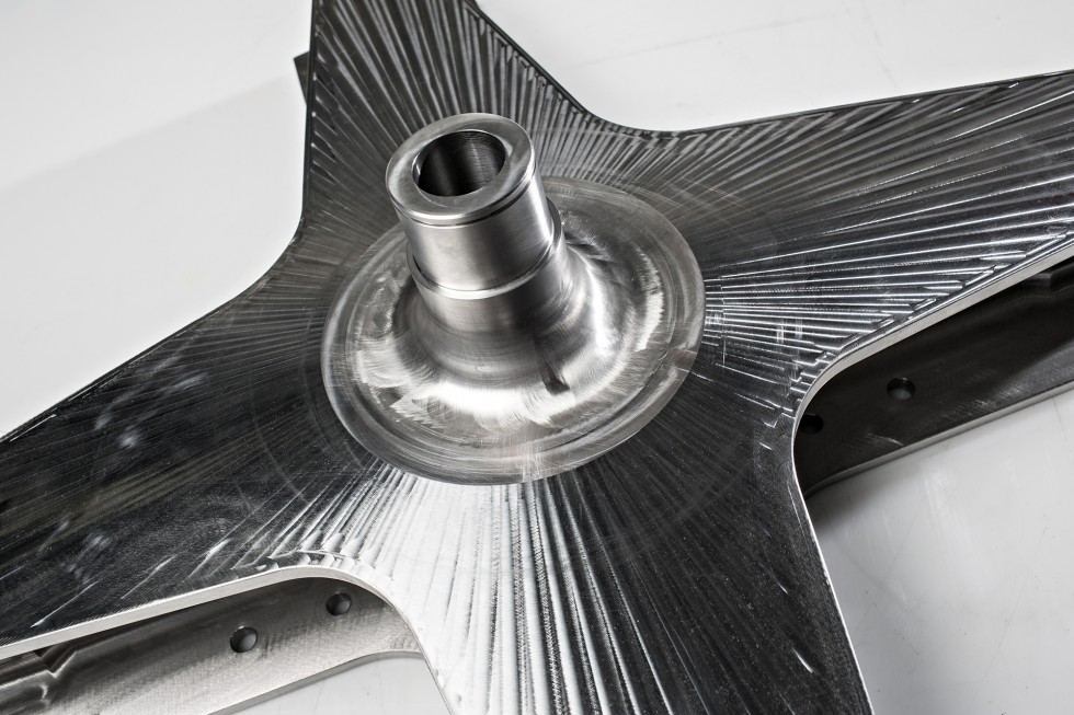 A metallic machined part resembling a four-point star with a spindle in the middle.